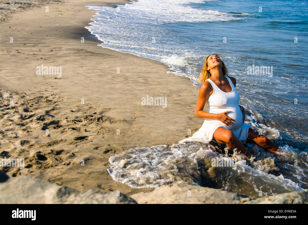 Pregnant mature woman sitting in ocean waves on beach whilst touching stomach Stock Photo