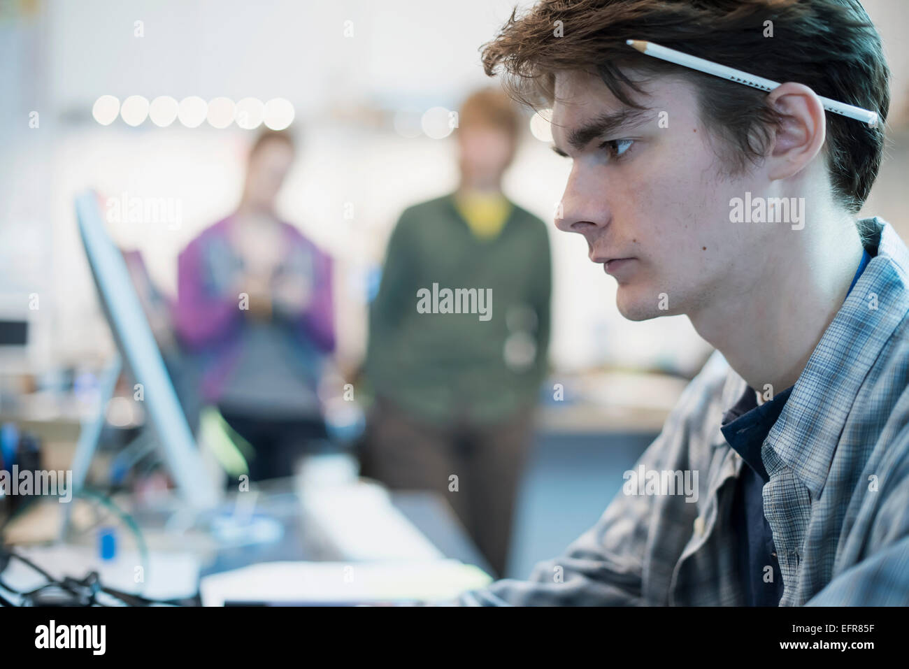 A young man seated at a computer in a repair shop.  Two people in the background. Stock Photo