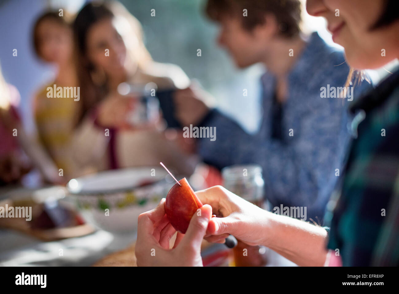 A group of people sitting at a table, eating and chatting. A woman slicing an apple. Stock Photo