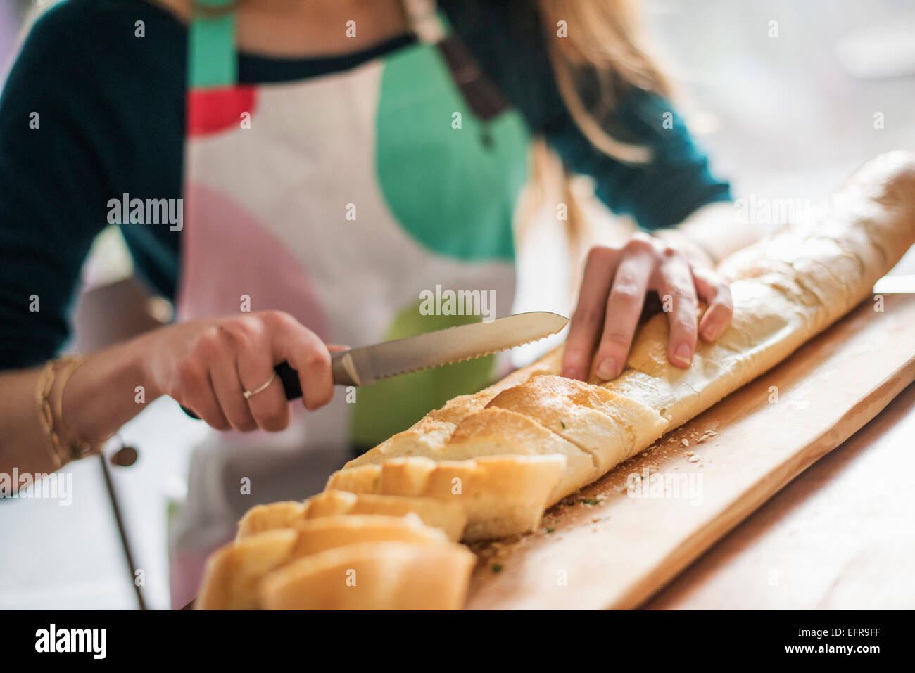 Close up of a woman wearing an apron, sitting at a table, slicing a baguette. Stock Photo