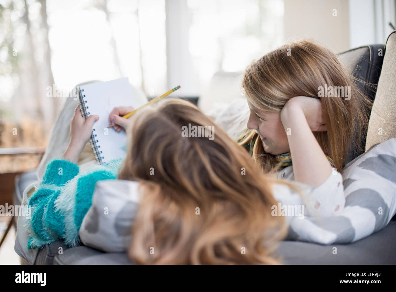 Two girls lying on a sofa, one writing into a notebook with a pencil. Stock Photo