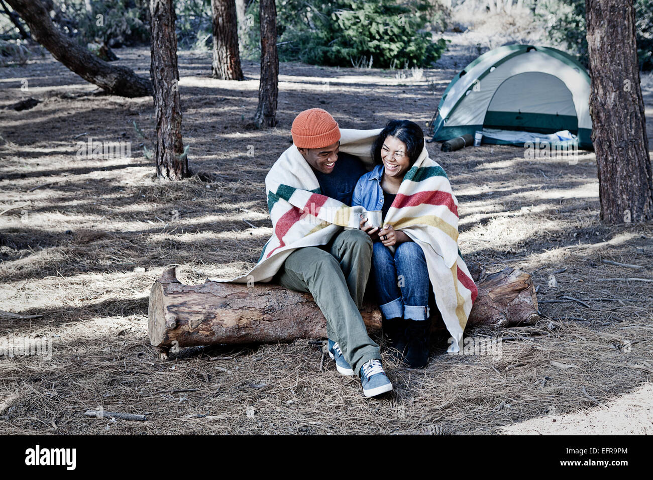 Young camping couple sitting on log wrapped in blanket Stock Photo