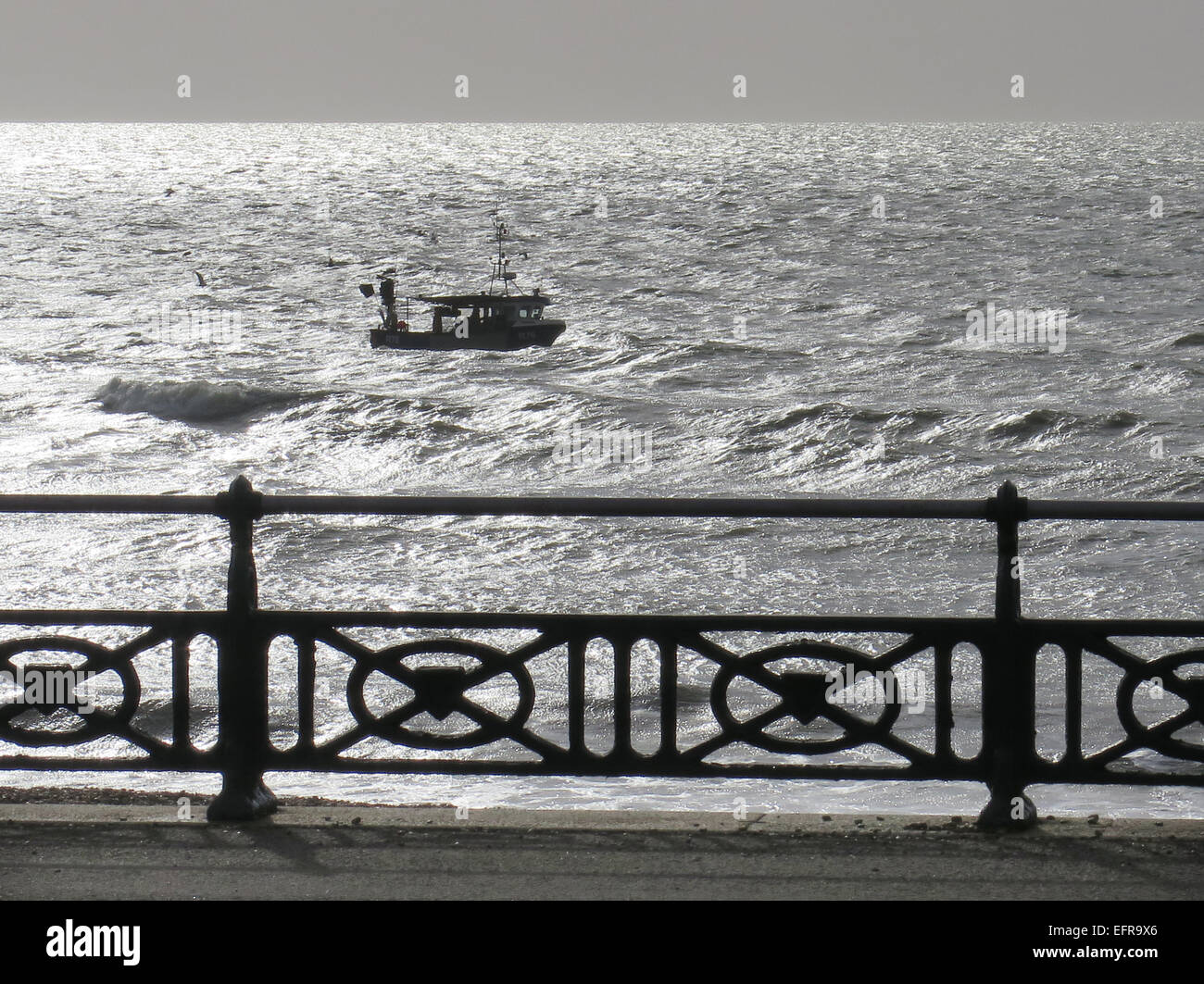 A fishing boat struggles against a westerly tide off Hove Lawns promenade in the early morning sunlight in the glistening sea. Stock Photo