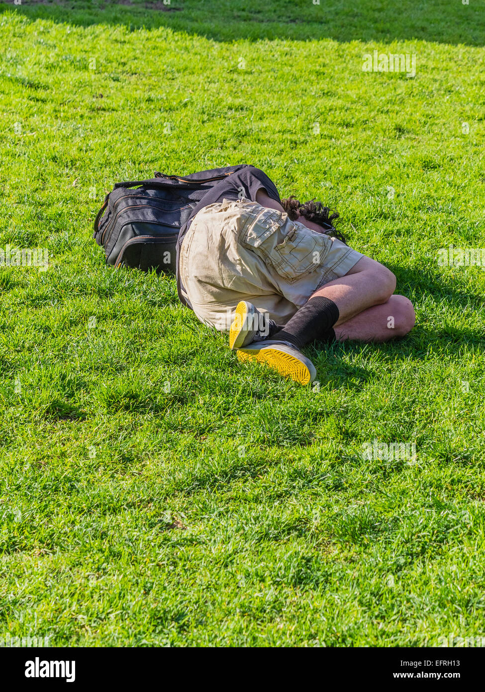 A young homeless man with bright yellow soles on his shoes sleeps lying down outside on the green grass. Stock Photo