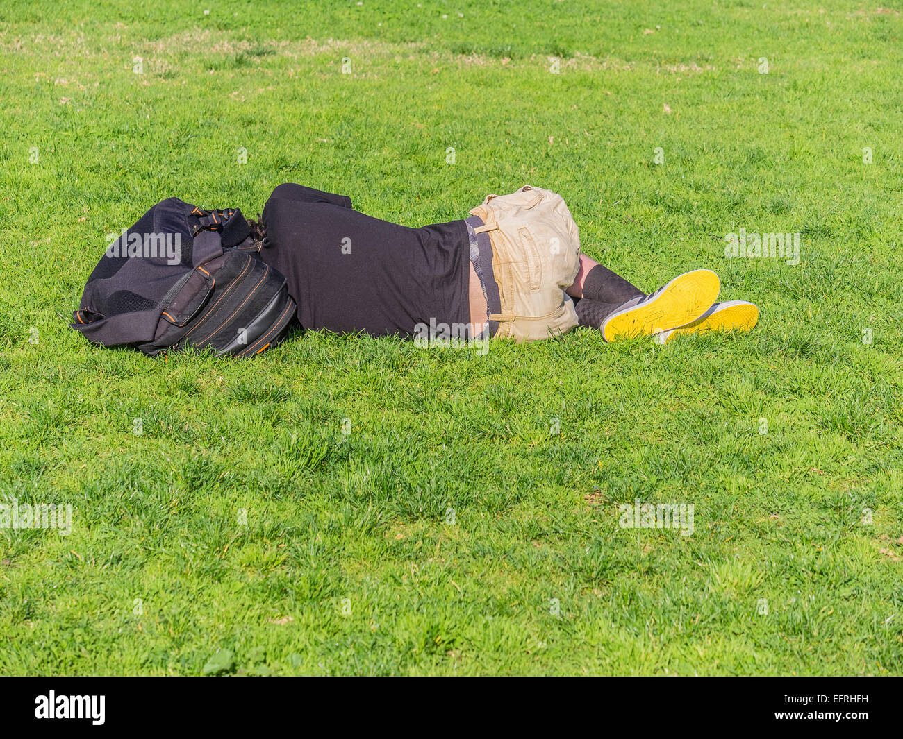 A young homeless man with bright yellow soles on his shoes sleeps lying down outside on the green grass. Stock Photo