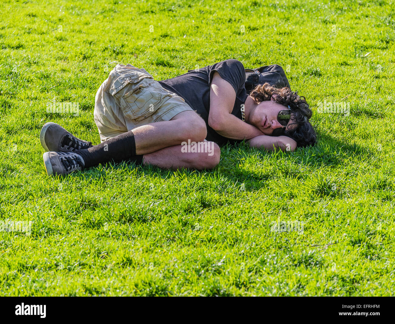 A young homeless man sleeps lying down outside on the green grass in