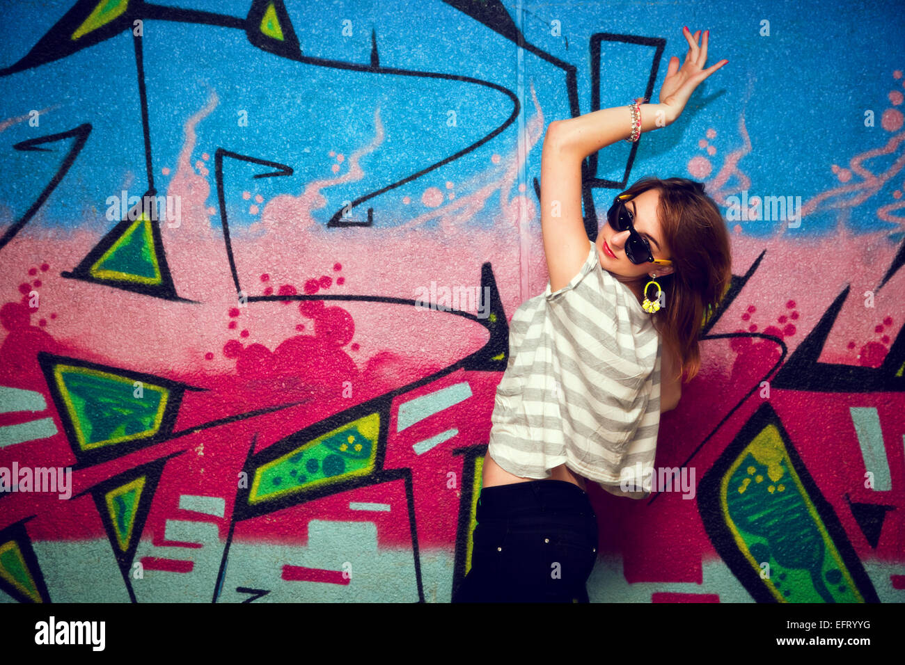 Stylish fashionable girl in a dance pose against colorful graffiti wall. Fashion, trends, subculture. Stock Photo