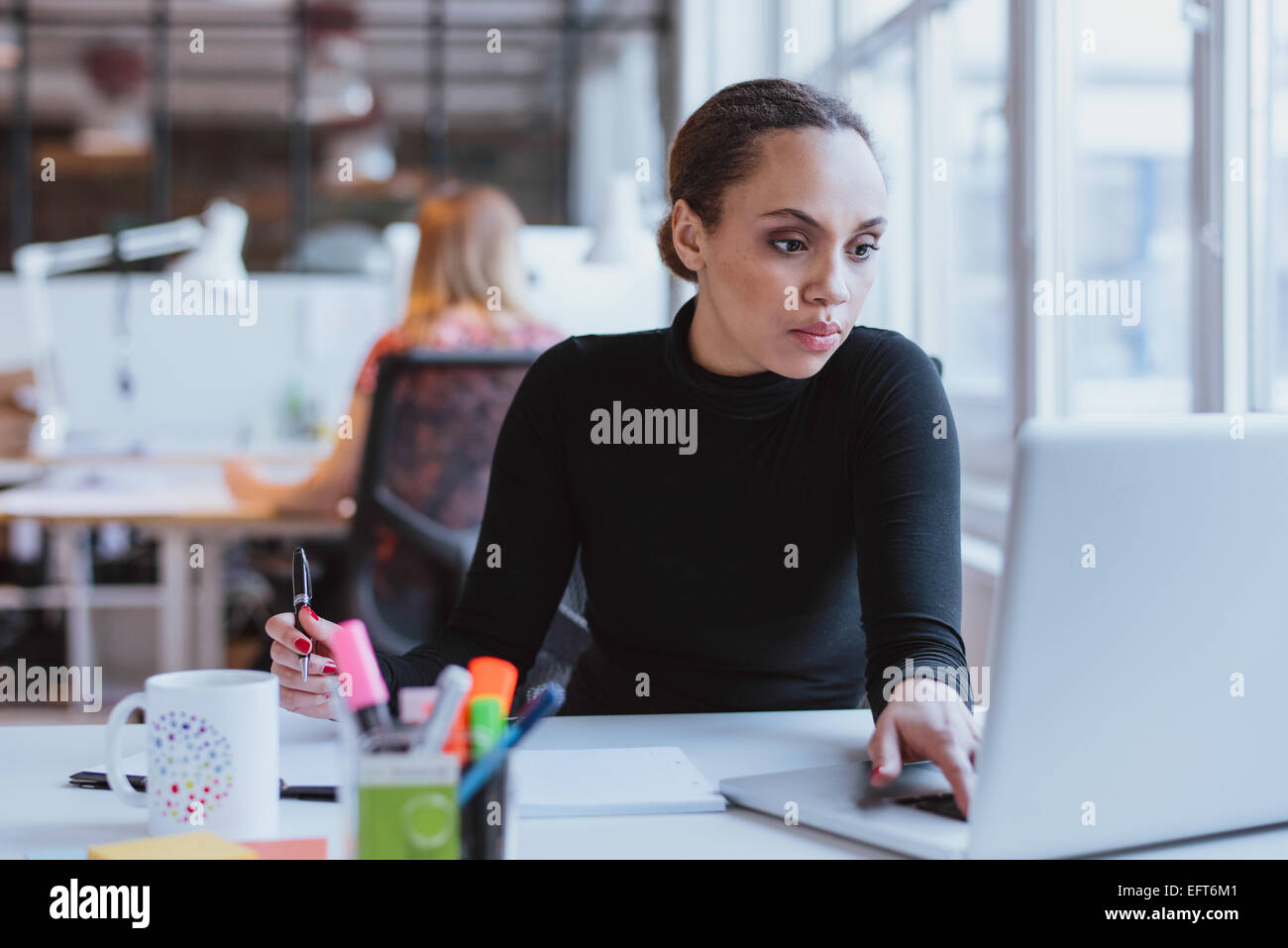 Image of young woman working on laptop while sitting at her desk in modern office. African female executive at work. Stock Photo