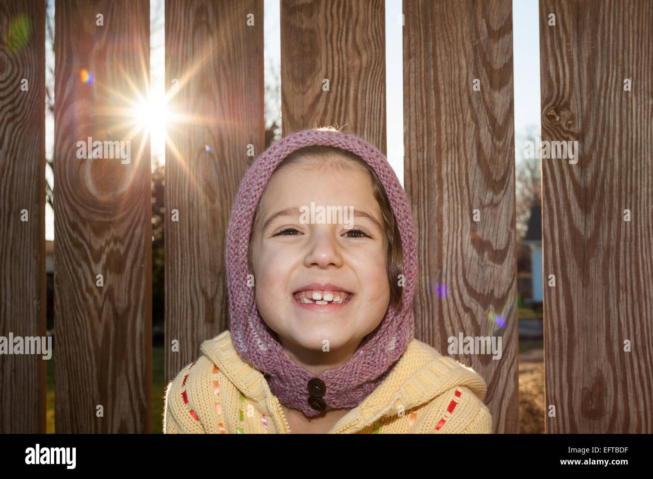 Young girl in knitted hat and sweater with a cute smile in front of a wooden fence. Stock Photo