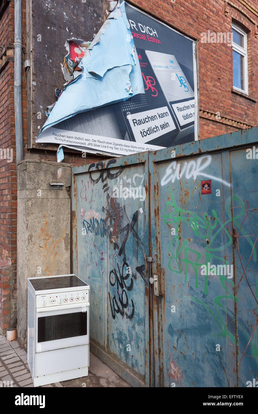 An old electric cooker sits in the street, next to a metal gate covered in graffiti and below a peeling advertising billboard Stock Photo