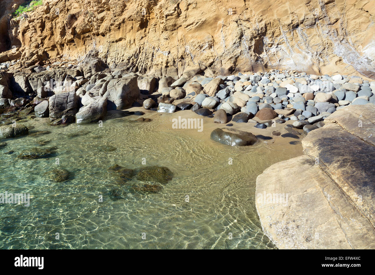 A small, secluded ocean front cove with large, smooth boulders embraces gentle surf bouncing off the large rocks during a bright Stock Photo