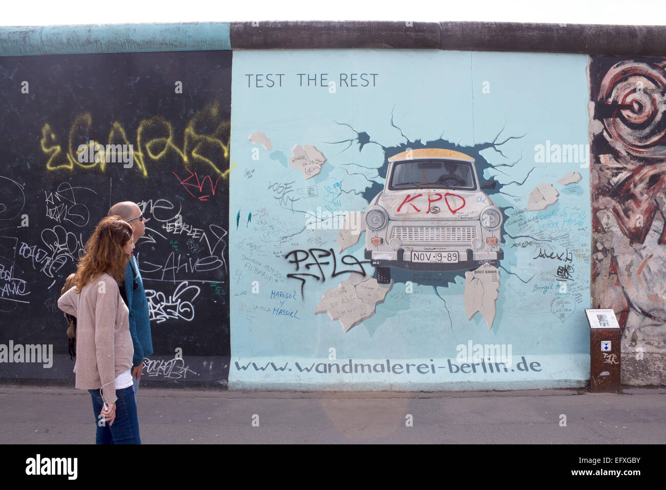 Couple walking past Birgit Kinder's artwork of Trabant car bursting through Wall Fifth update of artwork with additional graffit Stock Photo