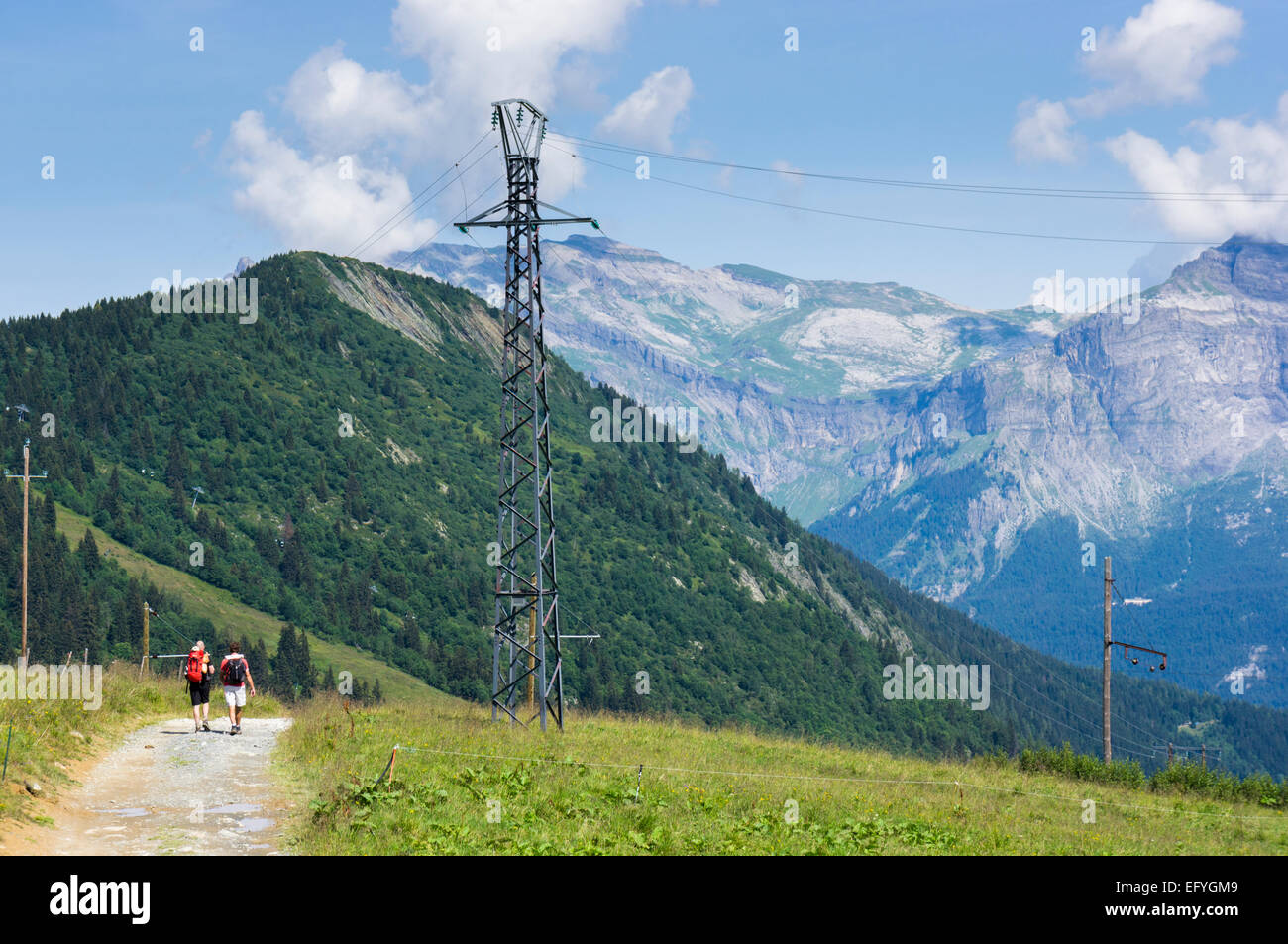 Walkers on path above in French Alps, Europe with high altitude electricity pylon taking power to remote mountain hotels / huts Stock Photo