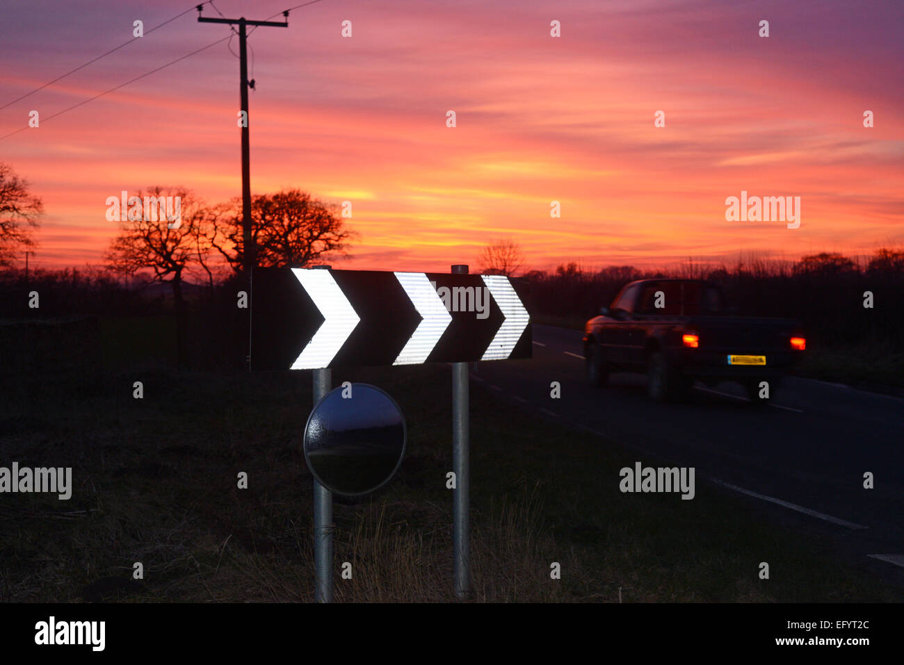 vehicle at sunset passing warning sign of sharp bend and circular safety mirror to give drivers view of road uk Stock Photo