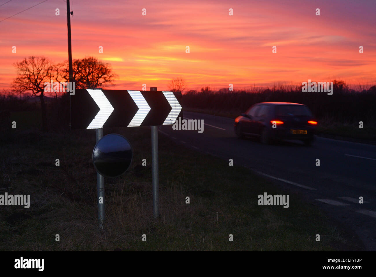 car at sunset passing warning sign of sharp bend and circular safety mirror to give drivers view of road uk Stock Photo