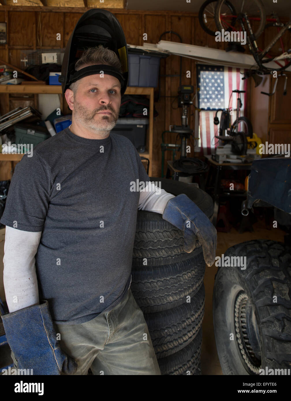Man wearing welding helmet leaning against tire with American flag in
