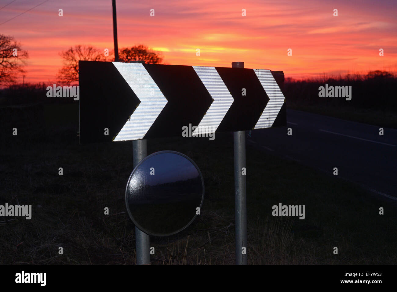 sunset by warning sign of sharp bend and circular safety mirror to give drivers view of road uk Stock Photo