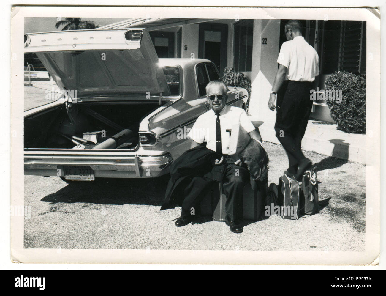 California, Usa. 12th Dec, 2014. CIRCA 1961: Reproduction of an antique photo shows businessman in white shirt and sunglasses holding a jacket, sitting on a suitcase near the open trunk of the car Dodge, the second man goes toward the motel, by car, in the background you can see the palm © Igor Golovniov/ZUMA Wire/Alamy Live News Stock Photo