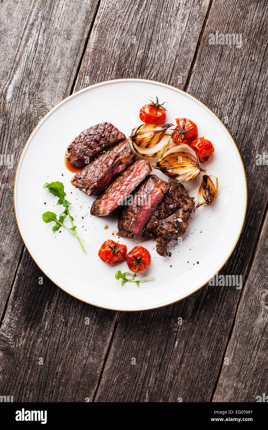 Sliced medium rare grilled Beef steak Ribeye with grilled onions and cherry tomatoes on plate on wooden background Stock Photo