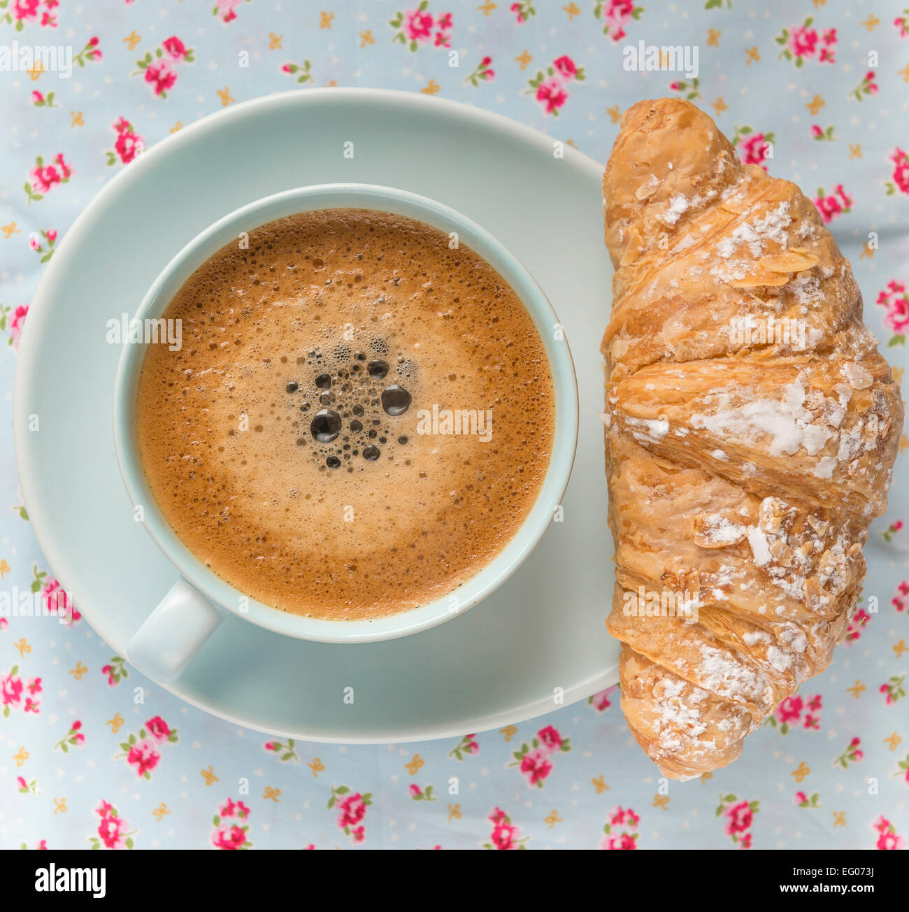Frothy coffee in a blue cup and saucer on a floral table cloth, with croissant pastry. Stock Photo