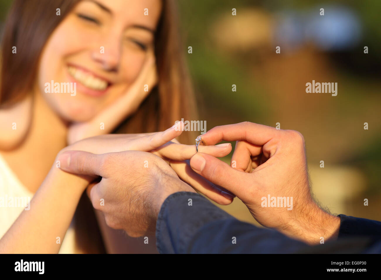 Boyfriend putting a engagement ring in his girlfriend finger outdoors with the smile of the girl in the background Stock Photo