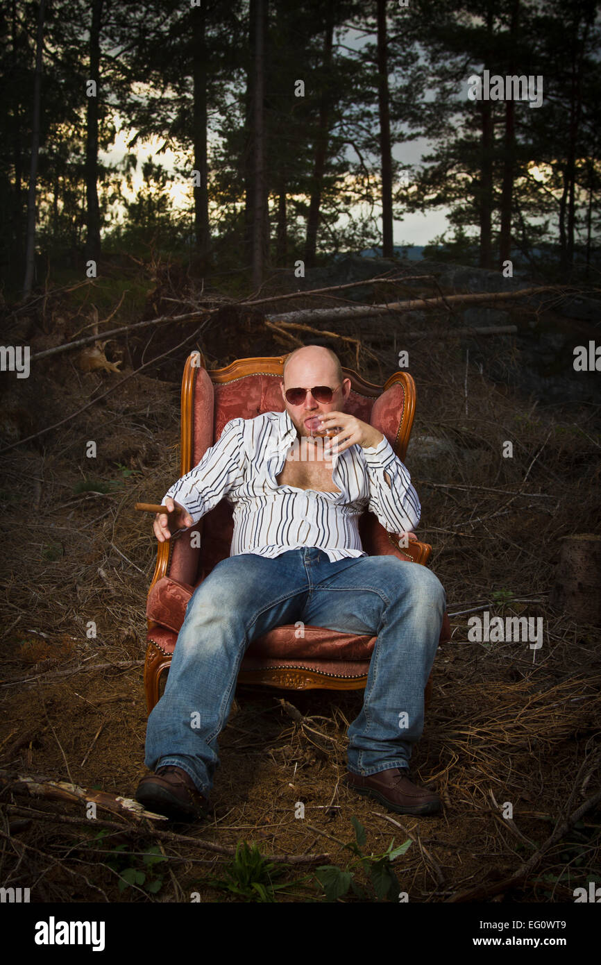 Sleazy man in a classic vintage chair in the middle of a logging area. Smoking cigar and drinking Cognac in woods. Stock Photo