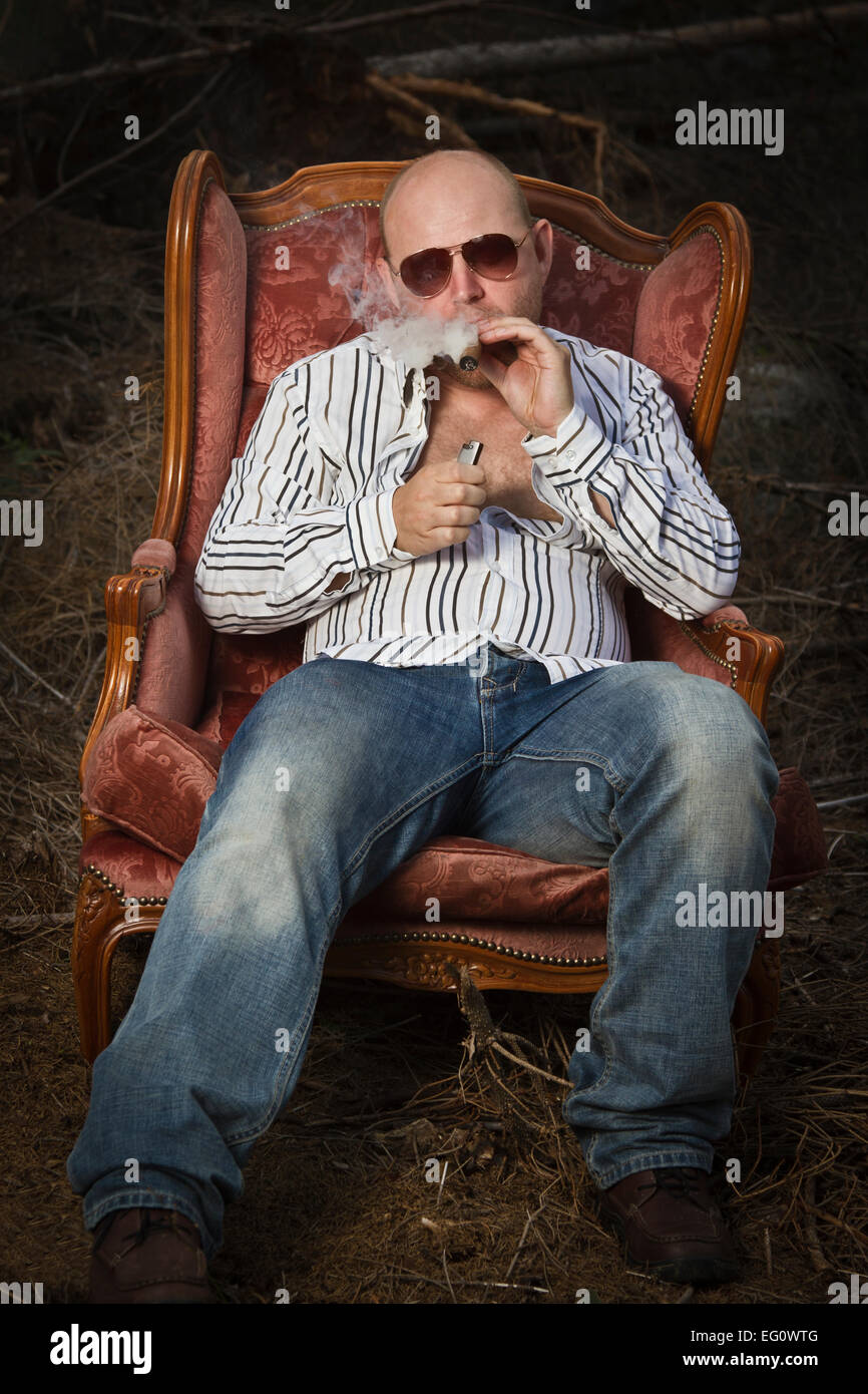 Sleazy man / dude in a classic vintage chair in the middle of a logging area. Fire up and smoke a cigar and drinking Cognac in w Stock Photo