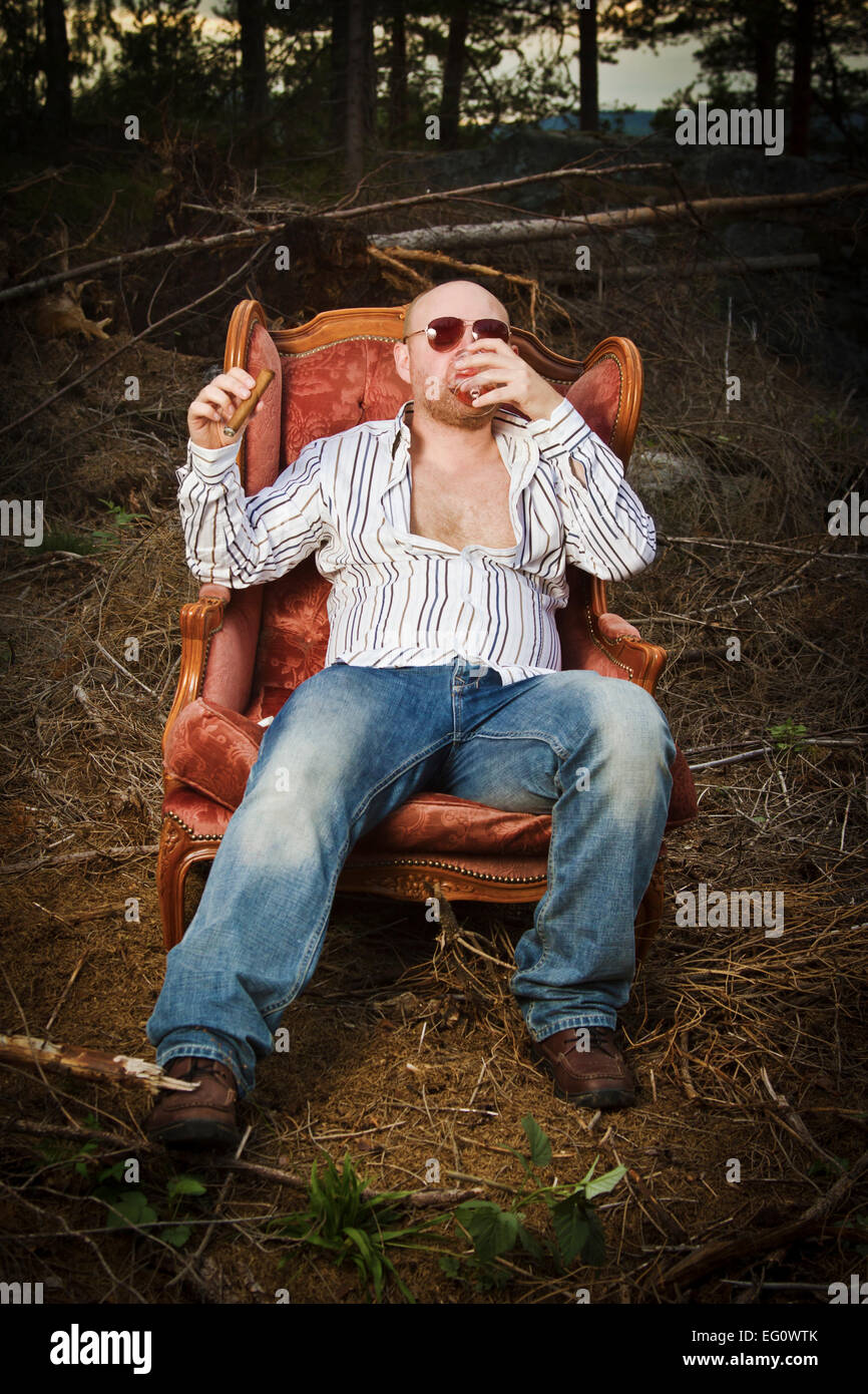 Sleazy man / dude in a classic vintage chair in the middle of a logging area. Smoke a cigar and drinking Cognac in woods. Stock Photo
