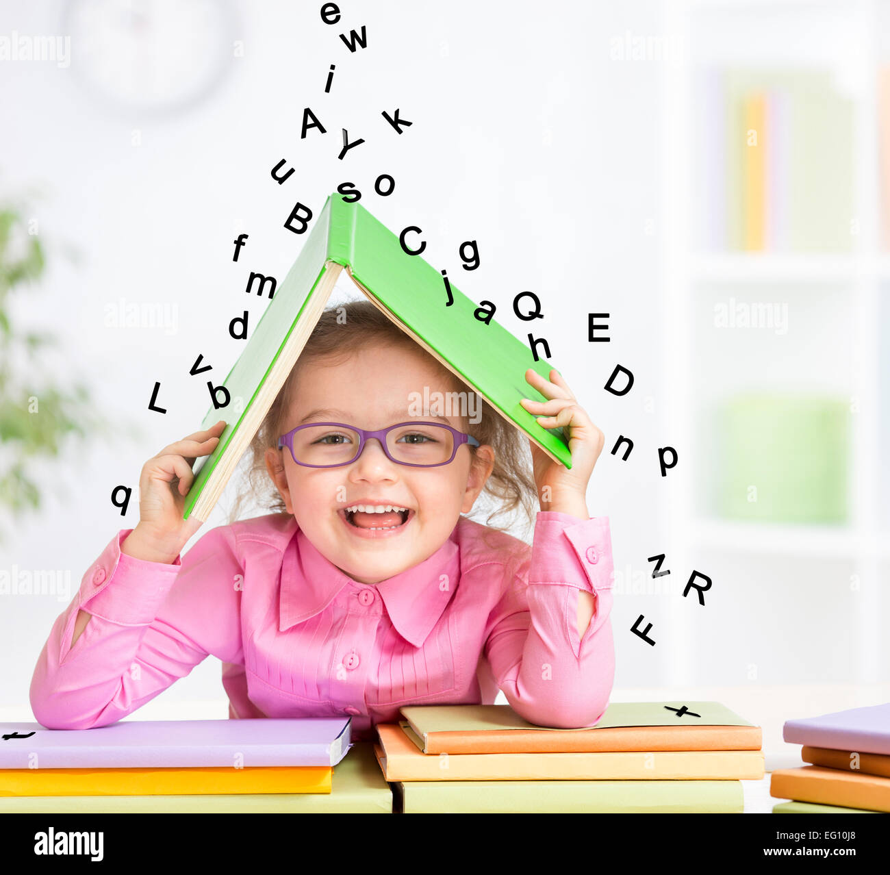 Smart smiling kid in glasses taking refuge under book roof from falling letters Stock Photo