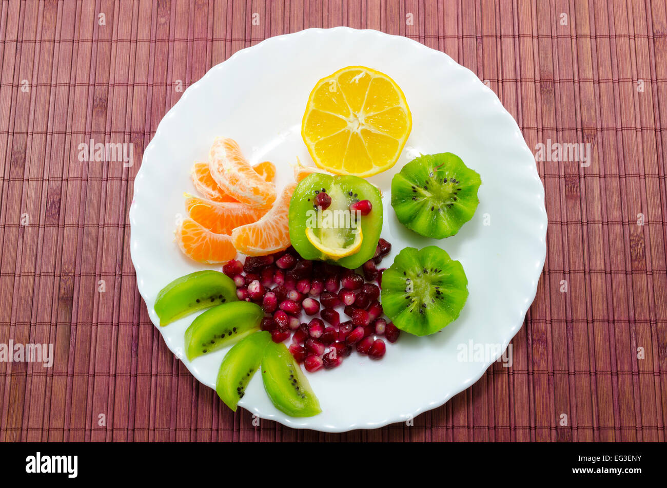 Sliced kiwi, pomegranate, oranges and lemon on a white plate, placed on a bamboo tablecloth Stock Photo