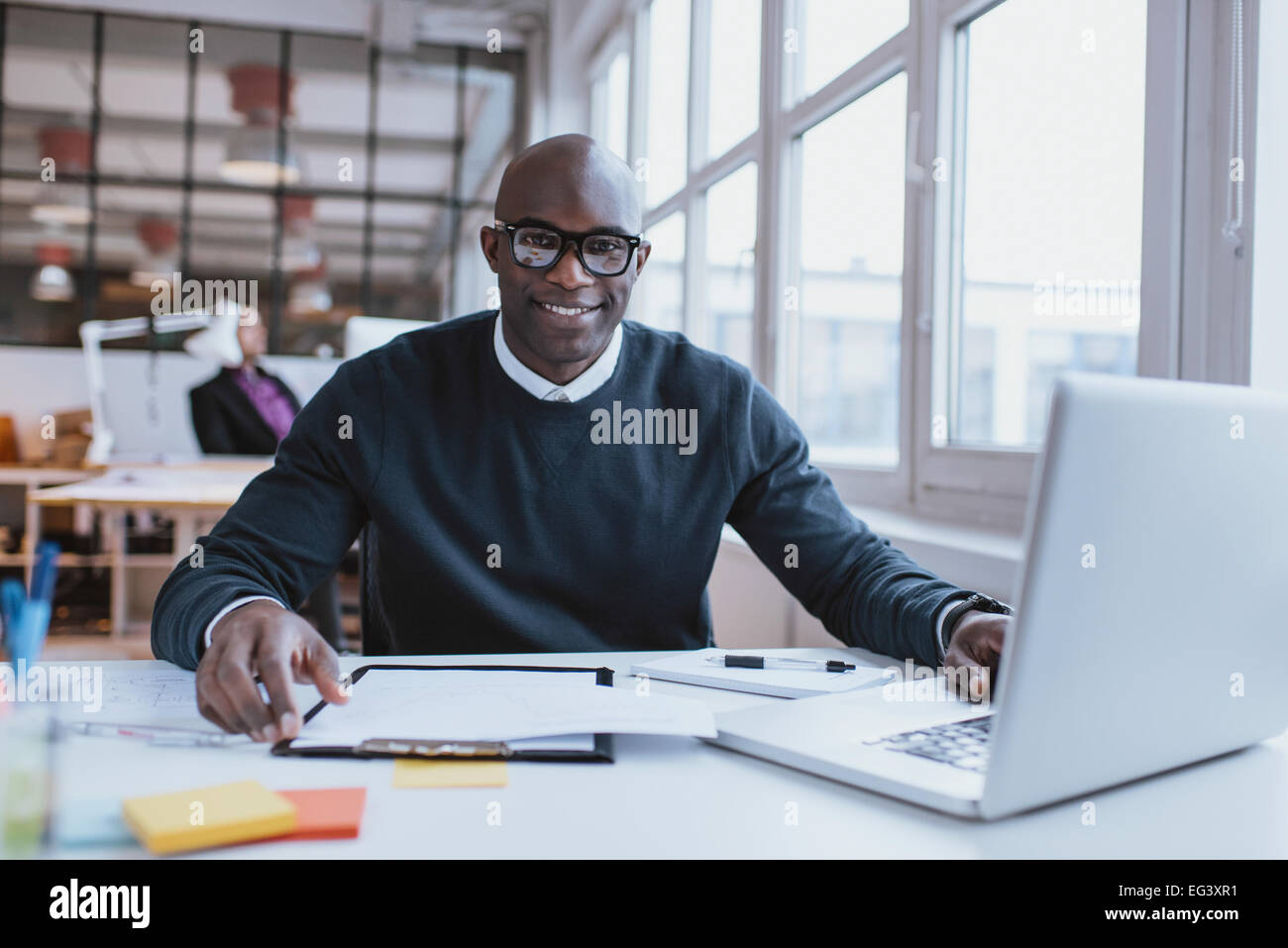 Portrait of confident young man at his desk with laptop doing paperwork. Happy african man looking at camera while at work. Stock Photo