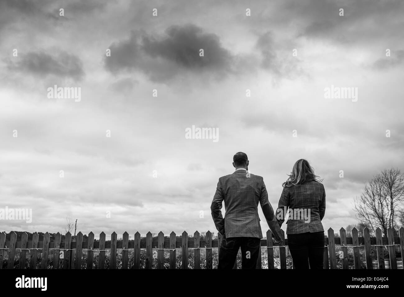 a black and white photograph of a man and woman holding hands standing against a wooden fence facing a stormy atmospheric sky Stock Photo
