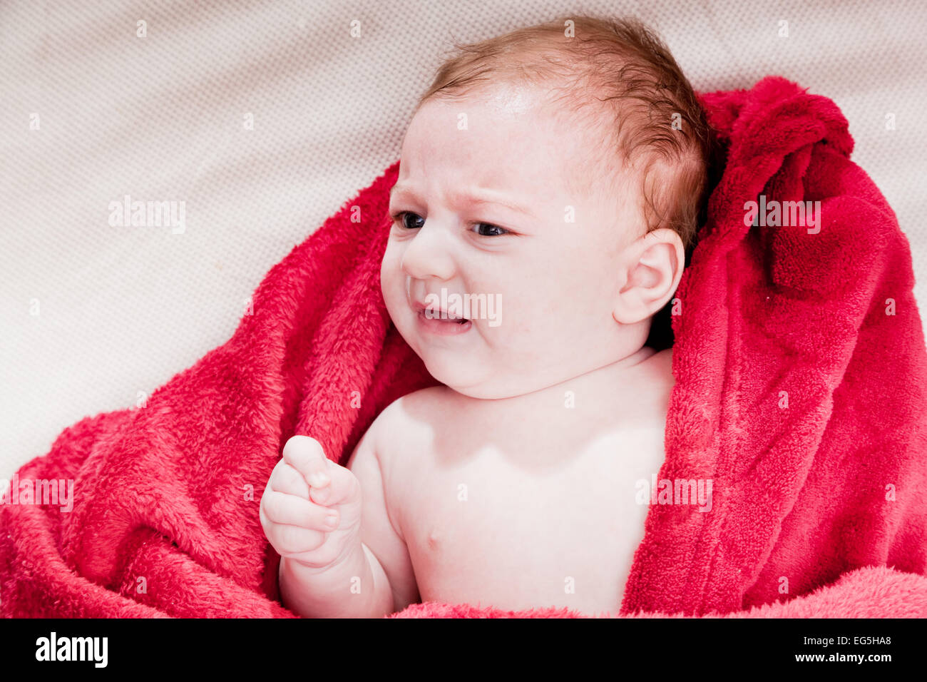 Lovely 3 months baby crying while lying on bed covered with red blanket