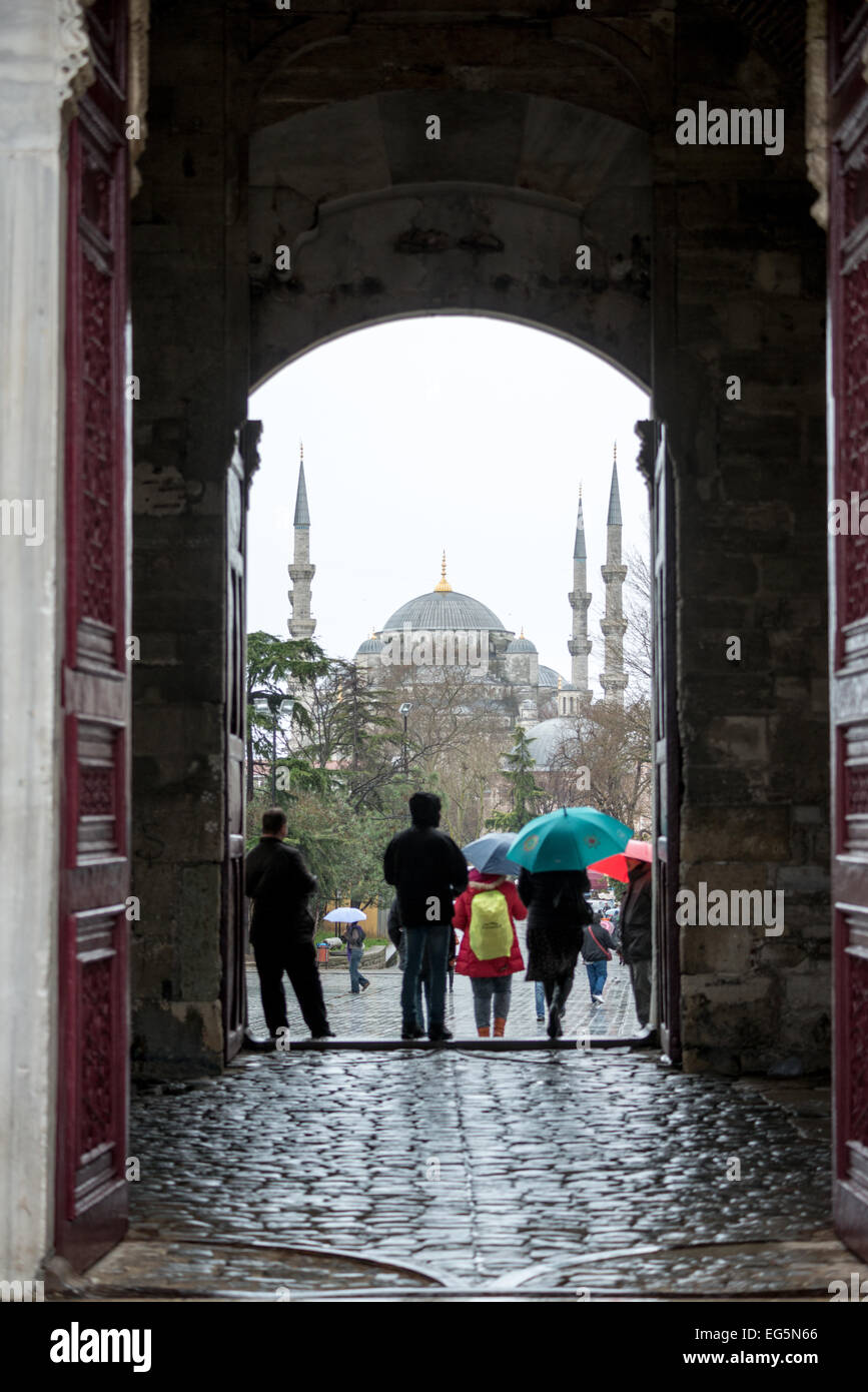 Visitors come and go in the rain through the Imperial Gate (Bâb-ı Hümâyûn) at Topkapi Palace, with tne entrance framing the Blue Mosque in the distance. On a peninsula overlooking both the Bosphorus Strait and the Golden Horn, Topkapi Palace was the primary residence of the Ottoman sultans for approximately 400 years (1465–1856) of their 624-year reign over Constantinople and the Ottoman Empire. Today it is one of Istanbul's primary tourist attractions. Stock Photo
