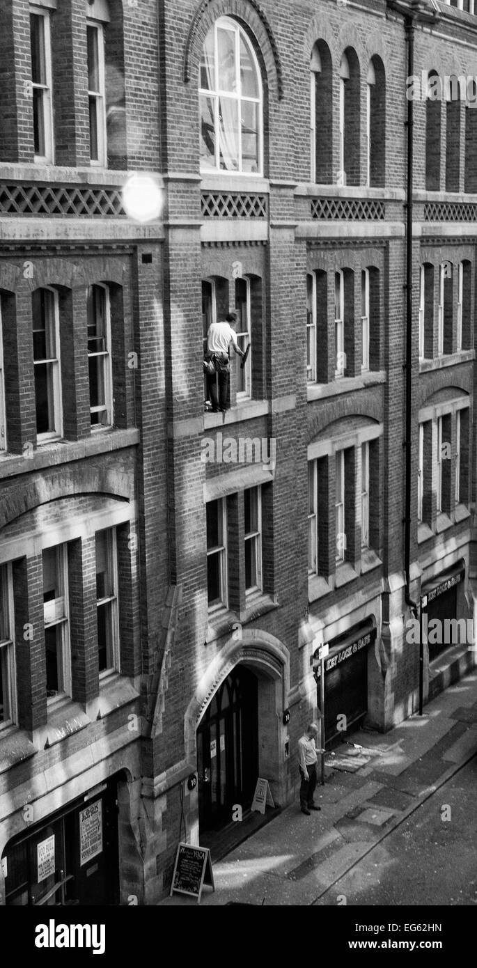 Window cleaner on very narrow ledge 3storeys up with no safety gear and man below oblivious to what is above him Stock Photo