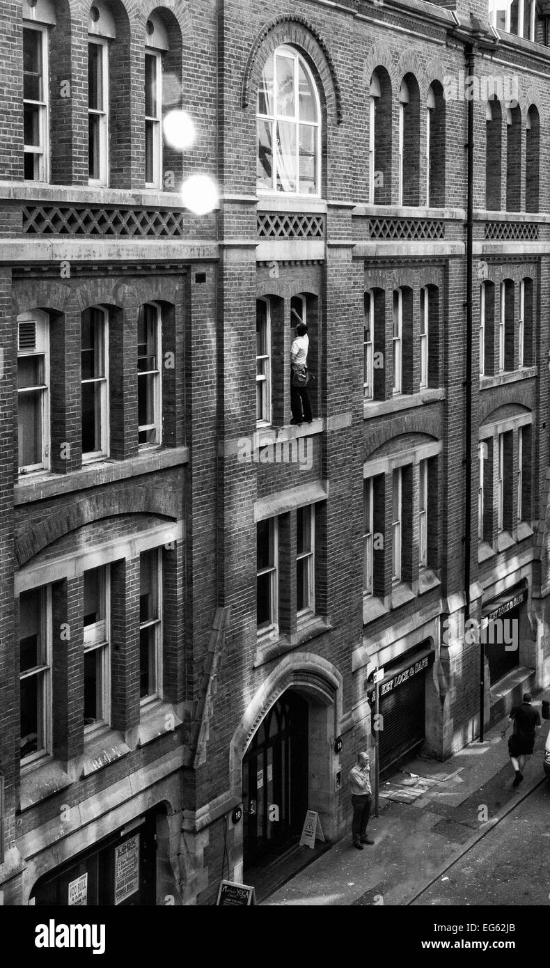 Window cleaner on very narrow ledge 3storeys up with no safety gear and man below oblivious to what is above him Stock Photo