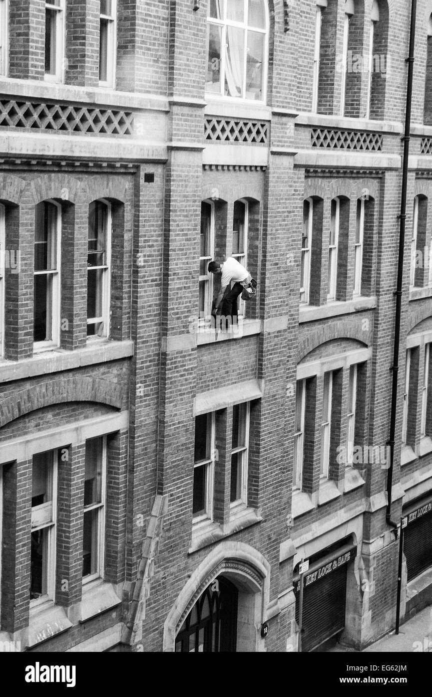 Window cleaner on very narrow ledge 3storeys up with no safety gear and man below oblivious to what is above him Stock Photo