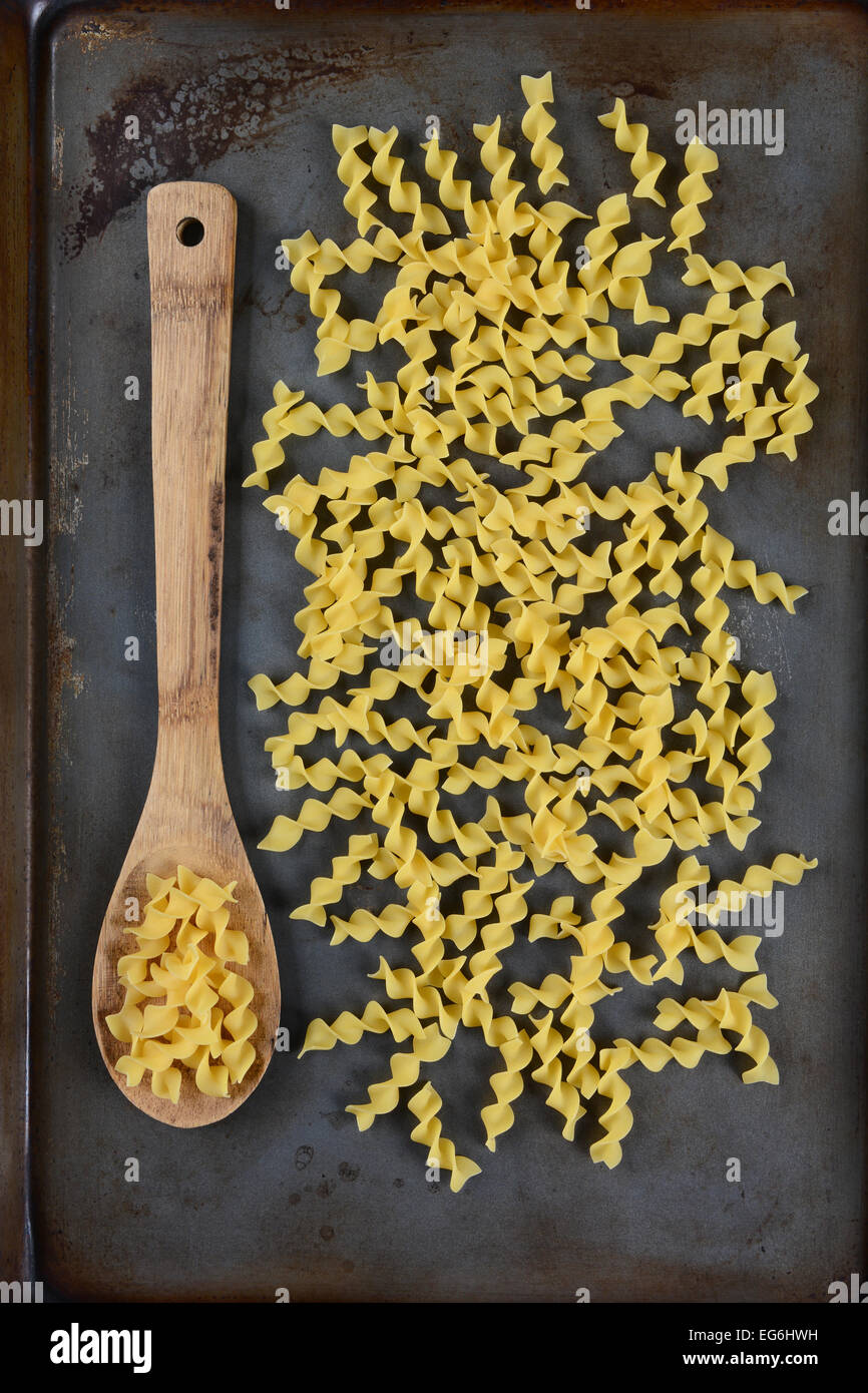 High angle shot of noodles scattered on a metal baking sheet with a wooden spoon holding some of the pasta. Vertical format. Stock Photo