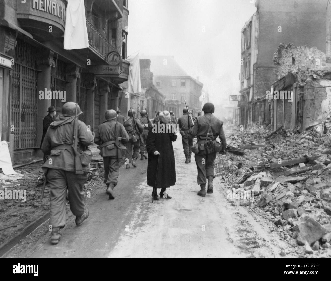 As infantrymen march through a German town, a shocked old woman stares at a the ruins. March-April 1945. Germany, World War 2. Stock Photo