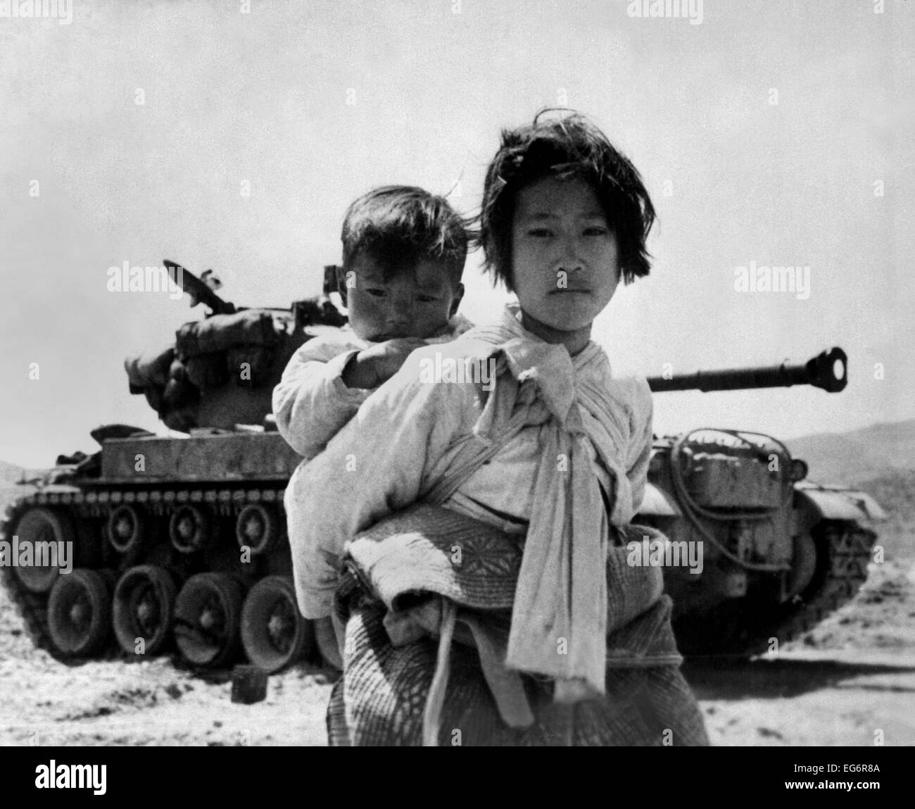 With her brother on her back, a war weary Korean girl tiredly trudges by a stalled M-26 tank, at Haengju, Korea. June 9, 1951. Stock Photo