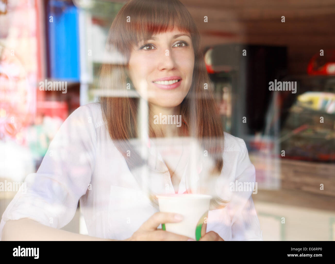 Young smiling woman behind glass of cafe. Pretty caucasian girl with cup of take away coffee Stock Photo