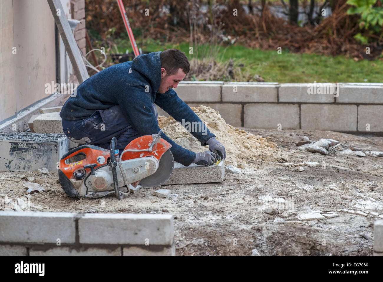 A young male laborer labourer bricklayer measures a cinder breeze block breezeblock CMU with a circular saw next to him. Stock Photo