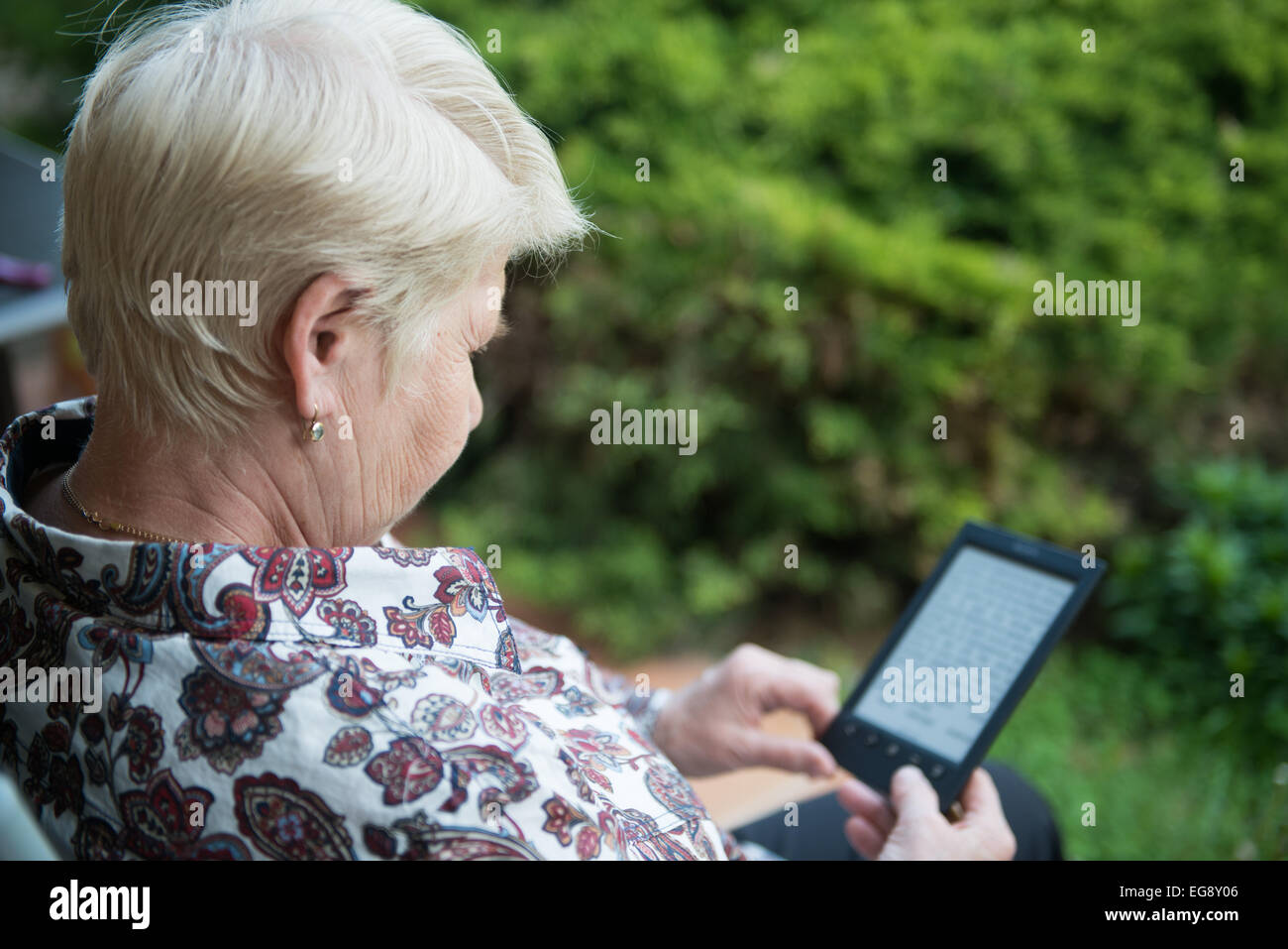 elderly senior woman sitting in the chair and reading e-book in the garden Stock Photo