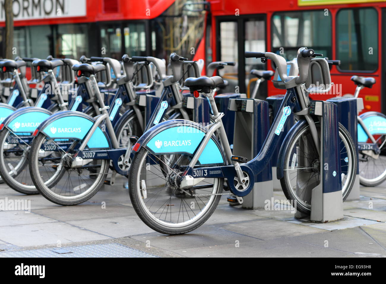 Blue Barclays Boris bikes line up on a street with red London buses behind Stock Photo