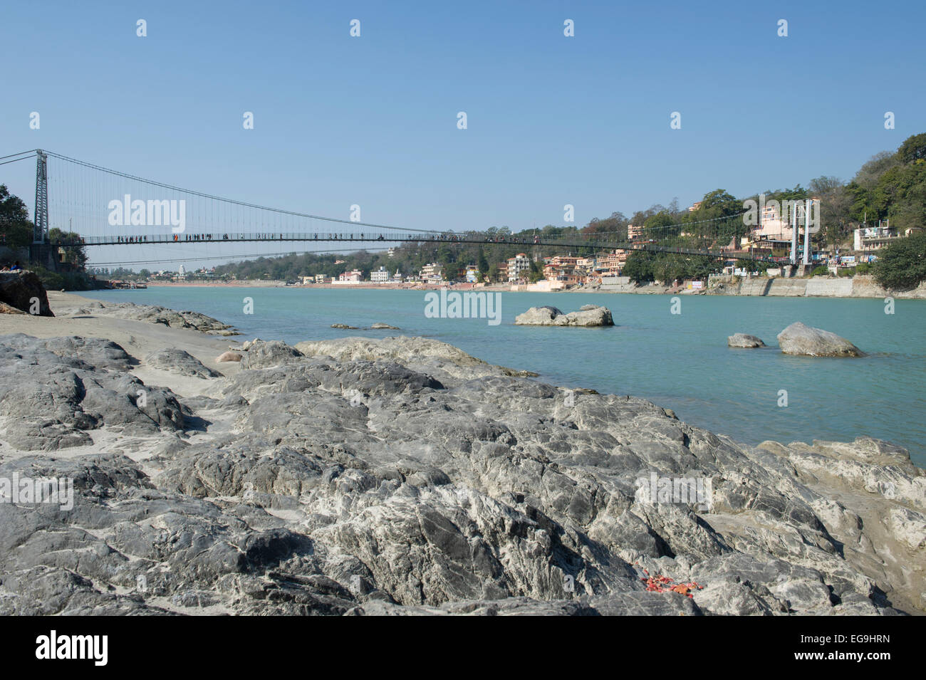 A rocky section of beach in Rishikesh with Ram Jhula bridge in the background which crosses the Ganges or Ganga river. Stock Photo
