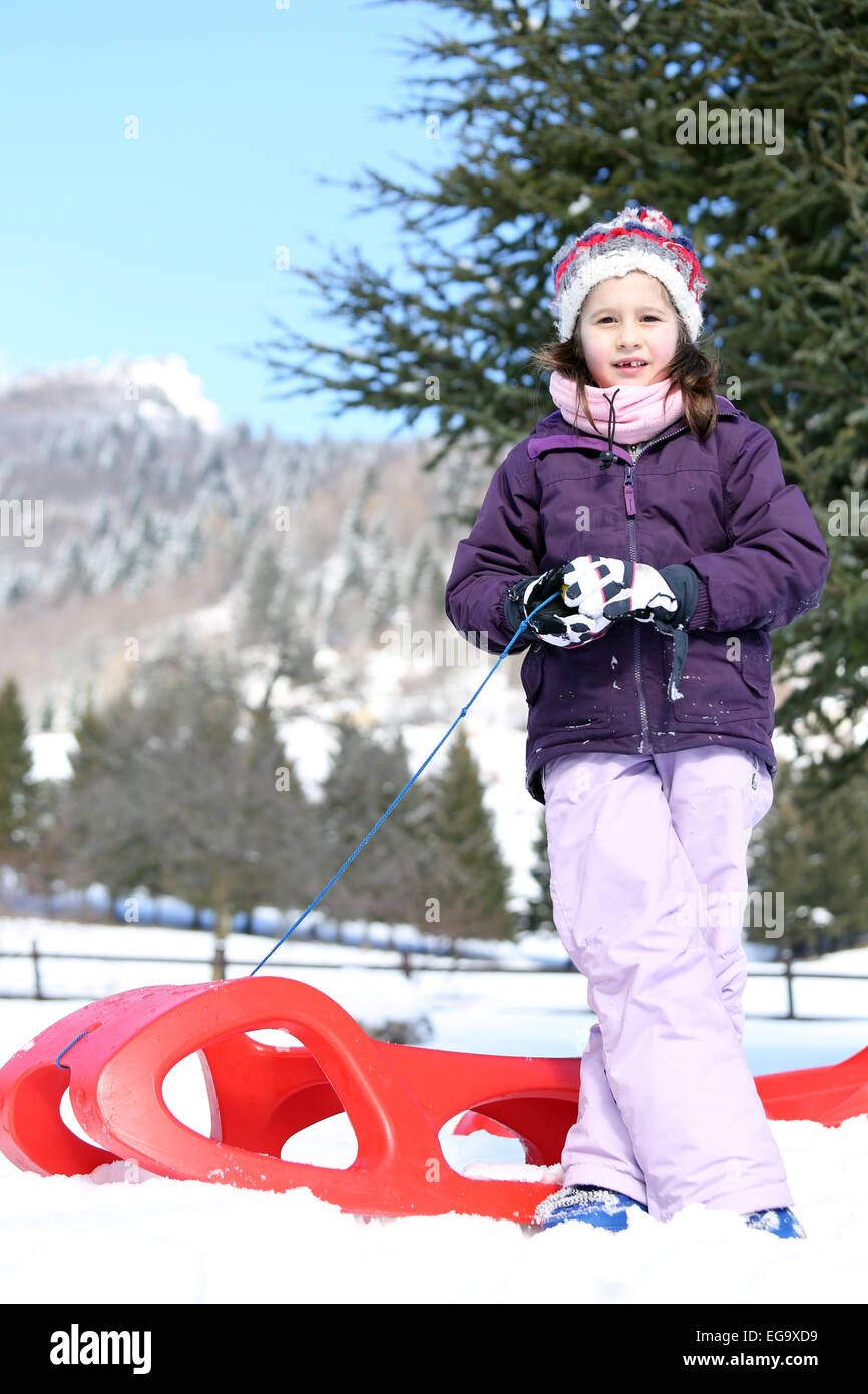 cute girl plays with the Red sled on the snow in the winter in the mountains Stock Photo