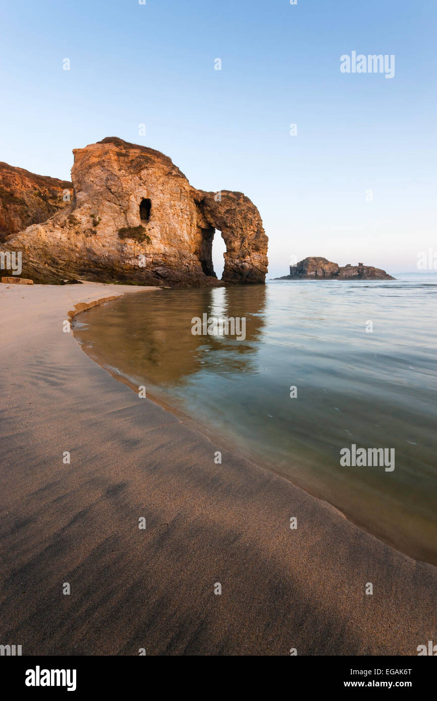 Perranporth arch is reflected in the sea on a clear morning in Cornwall. Textured sand sweeps around the sea towards the arch. Stock Photo