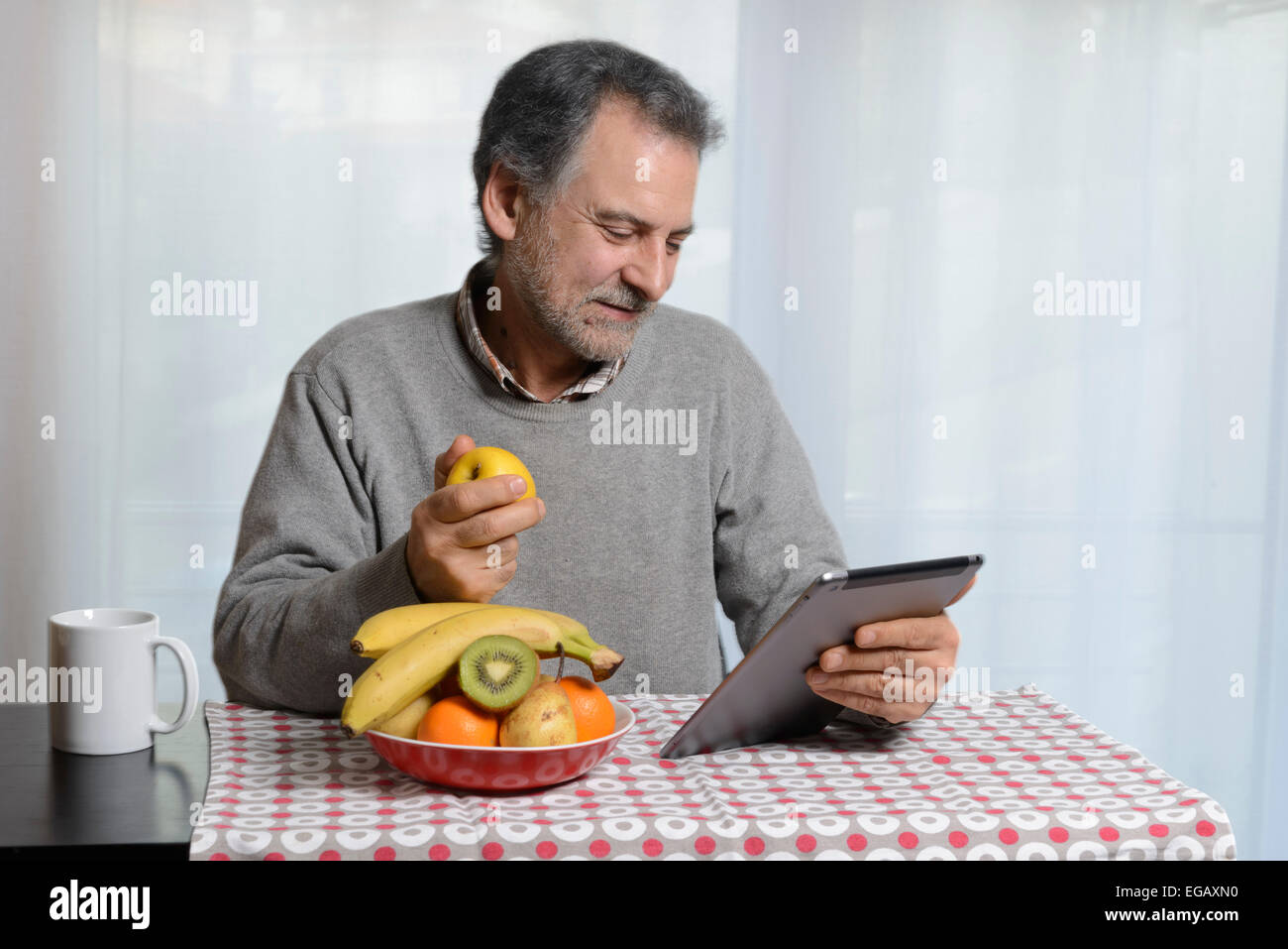 Middle aged man using an Apple iPad while eating fruit for breakfast in the kitchen at home Stock Photo