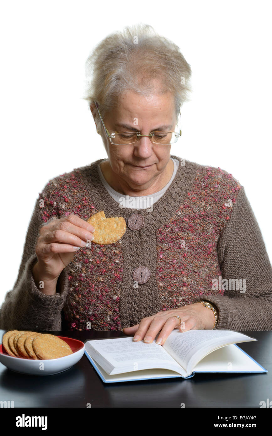 Middle aged woman reading a book while eating cookies at home Stock Photo