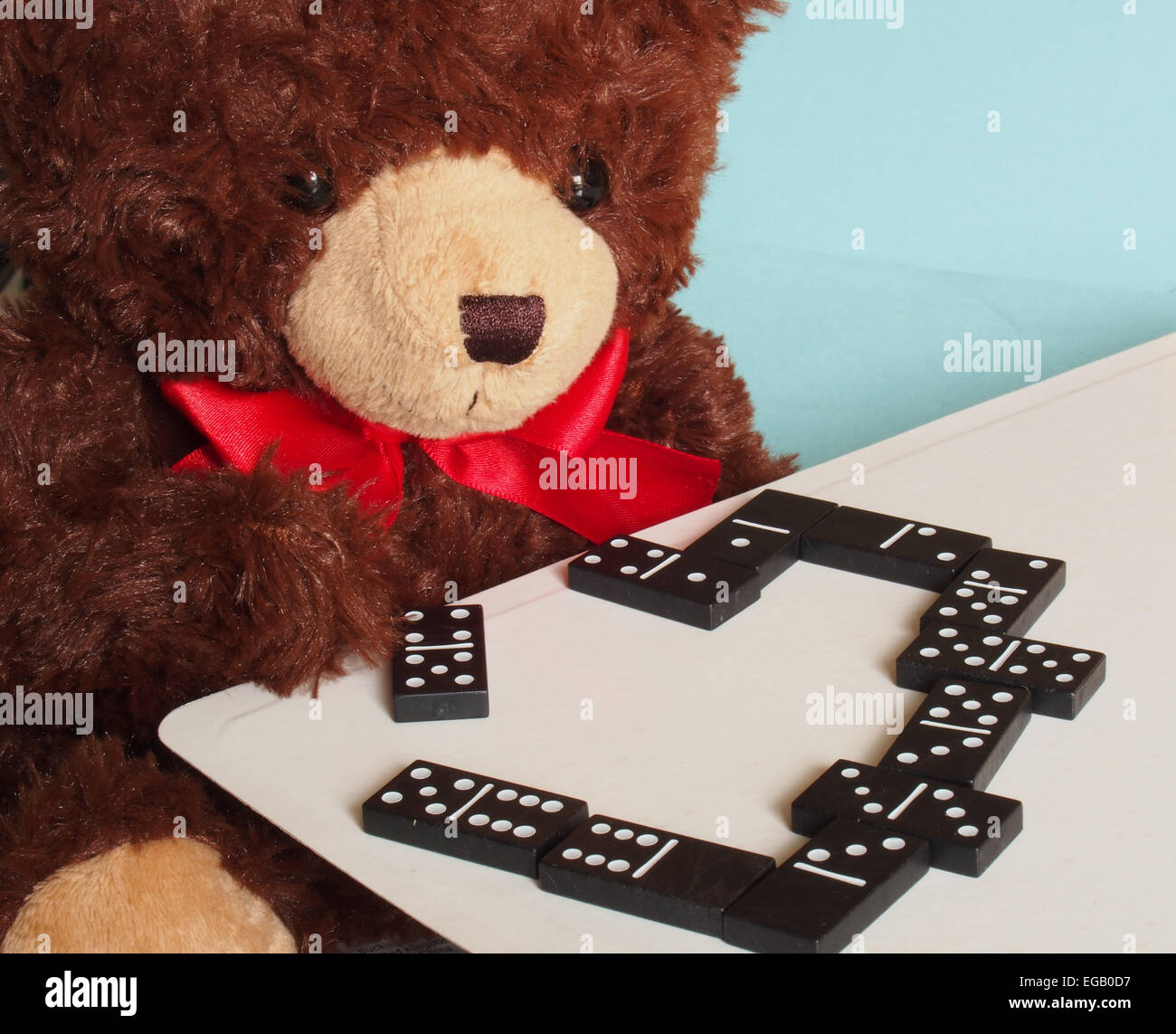 A soft brown teddy bear playing a game of dominoes and concentrating hard wearing a big red ribbon bow Stock Photo